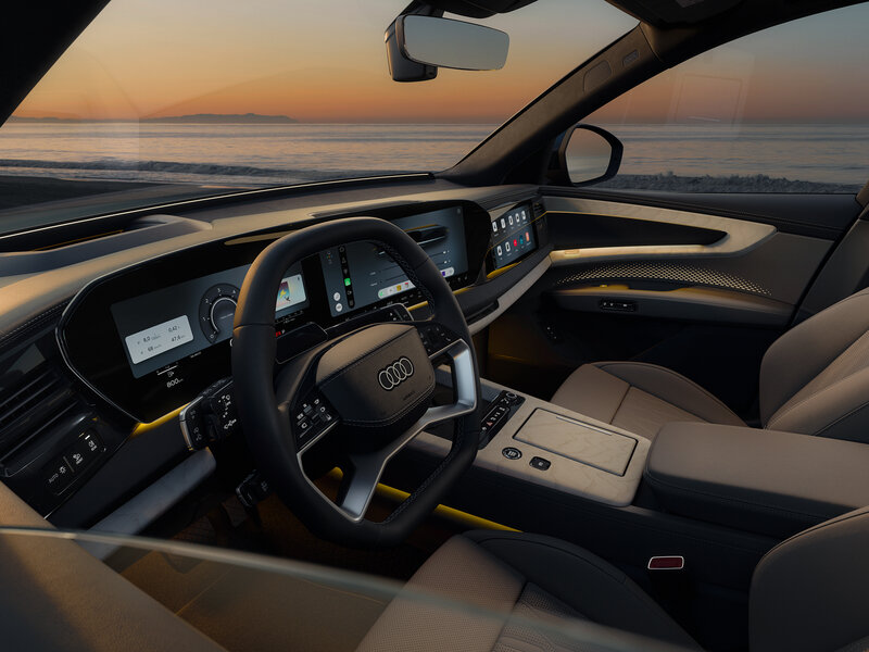 Cockpit des Audi Q9 mit digitalem Kombiinstrument, großem Zentraldisplay und hellem Interieur mit Blick über das Armaturenbrett zur Straße bei Sonnenuntergang