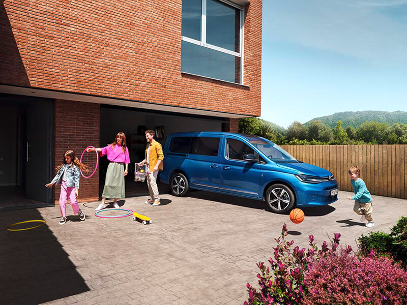 Ein blauer Volkswagen Caddy California steht vor einem Wohnhaus mit geöffneter Schiebetür. Mehrere Personen, darunter Kinder, bewegen sich um das Fahrzeug. Der Fokus liegt auf Alltagstauglichkeit und Familiennutzung.