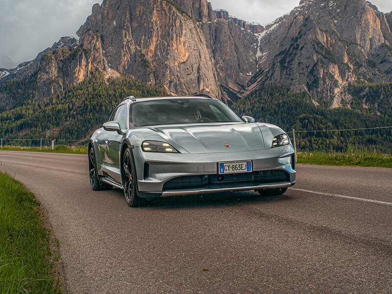 Ein elektrischer Porsche Taycan Cross Turismo fährt auf einer Landstraße vor alpiner Bergkulisse.