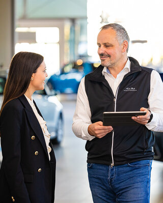 Beratungssituation im Autohaus: Ein Mitarbeiter zeigt einer Kundin Fahrzeugdetails auf einem Tablet, umgeben von ausgestellten Pkw.