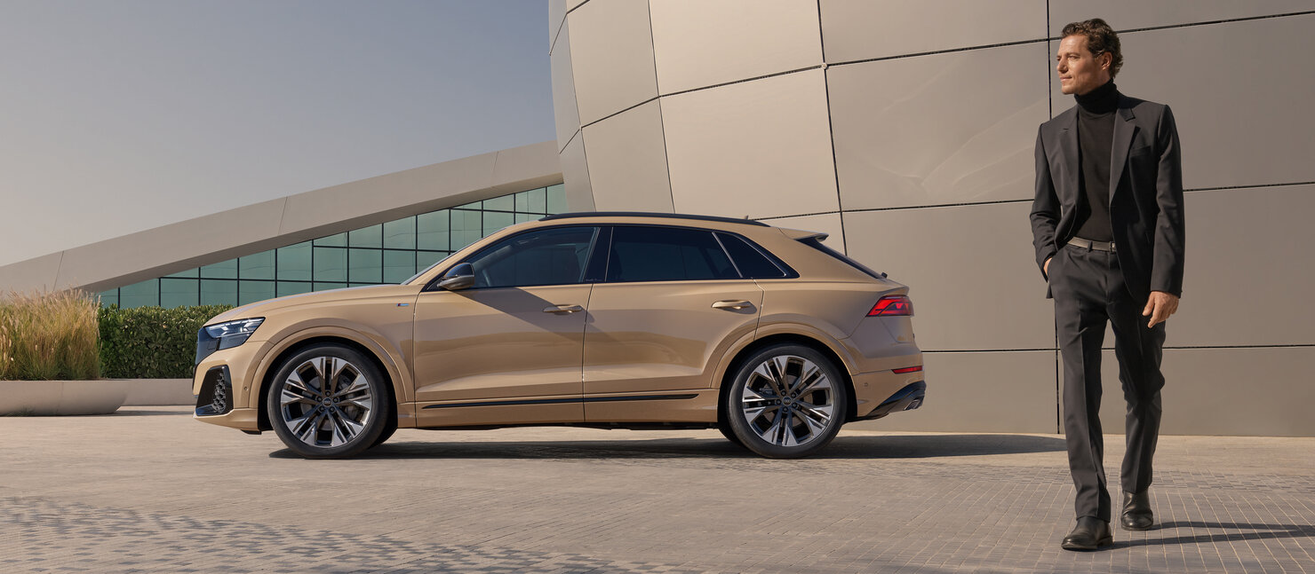 Audi Q8 in Seitenansicht vor moderner Fassade, elegante Linienführung des SUVs mit Person im Vordergrund als Größenvergleich