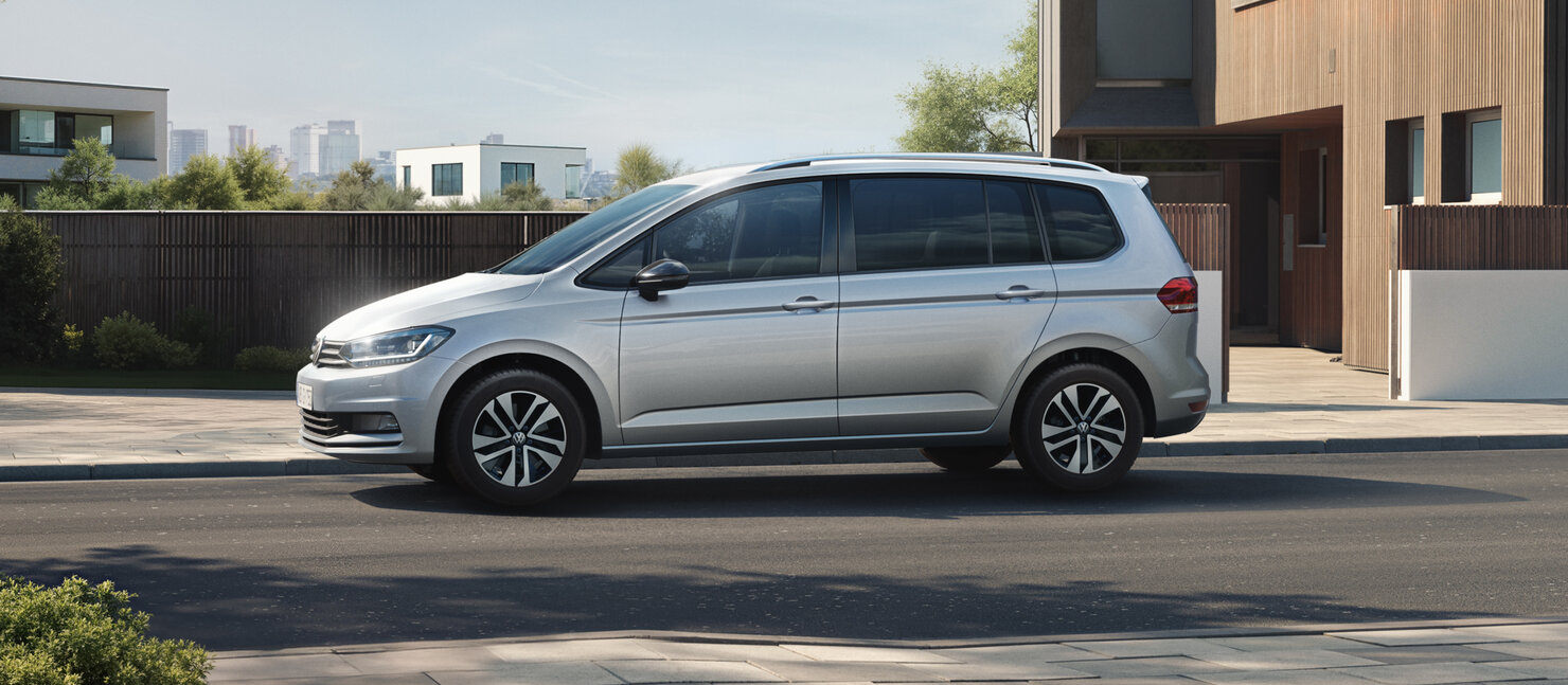 Volkswagen Touran in Silber als Seitenansicht vor moderner Wohnarchitektur, betont großzügigen Innenraum und Alltagstauglichkeit