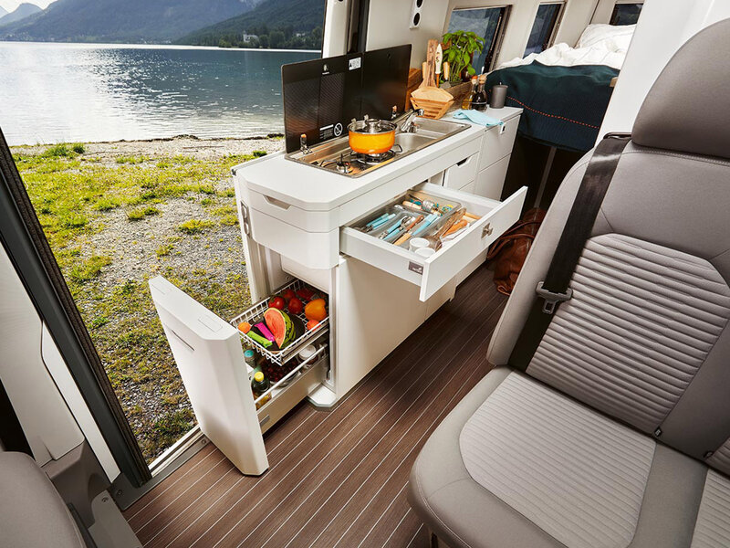 Innenansicht des Volkswagen Grand California mit integrierter Küchenzeile, ausgezogenen Schubladen und sichtbarem Stauraum. Blick nach draußen auf eine natürliche Landschaft.