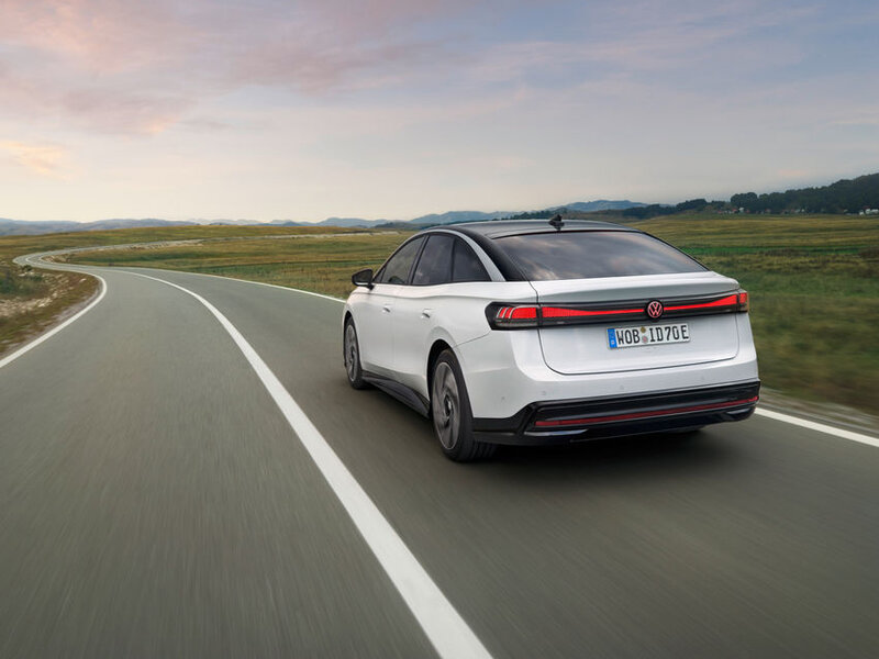 Weißer Volkswagen ID.7 fährt auf einer Landstraße durch offene Landschaft und ist von hinten schräg zu sehen.