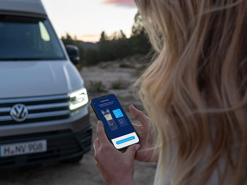 Eine Person hält ein Smartphone vor einem geparkten Volkswagen Grand California und steuert Fahrzeugfunktionen über eine App.