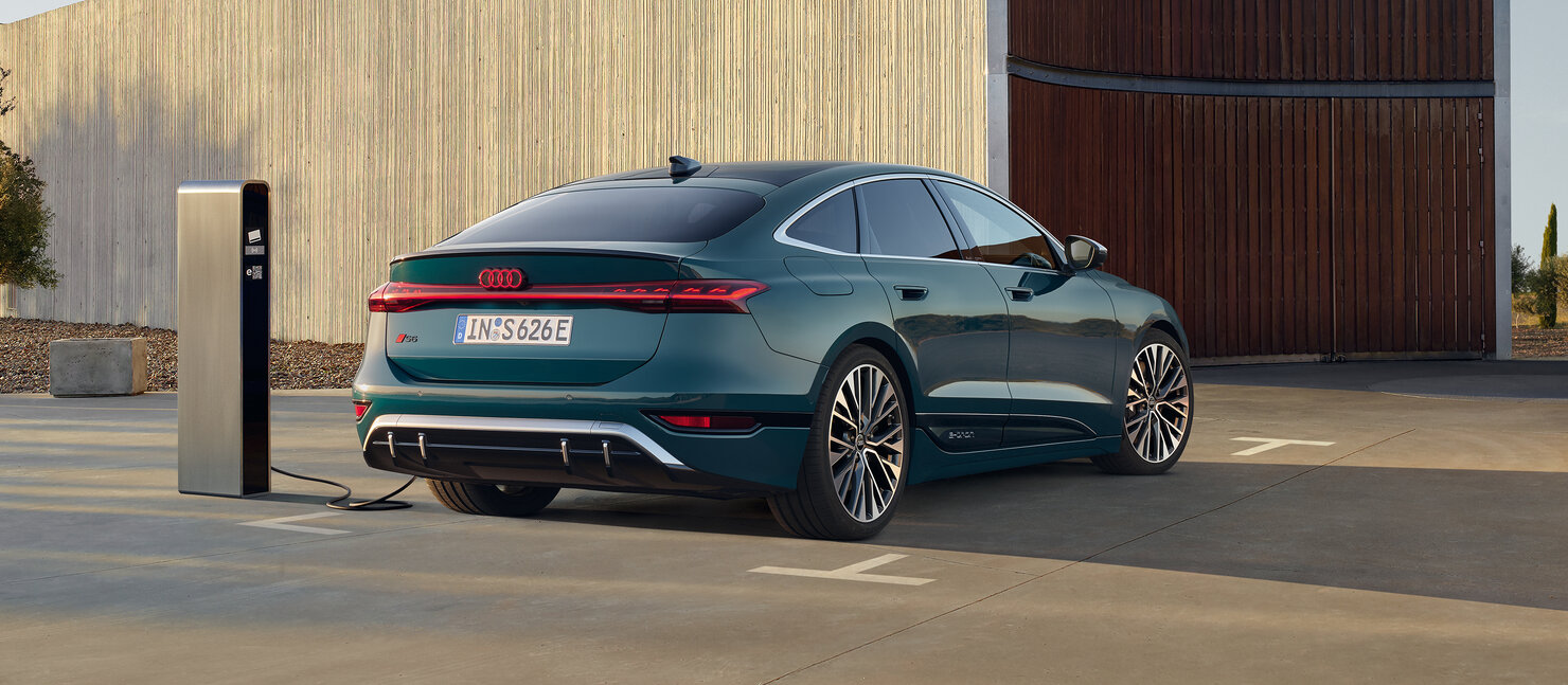 Audi S6 Sportback e-tron an Schnellladesäule vor moderner Architektur mit coupéhafter Dachlinie und breitem Heckauftritt