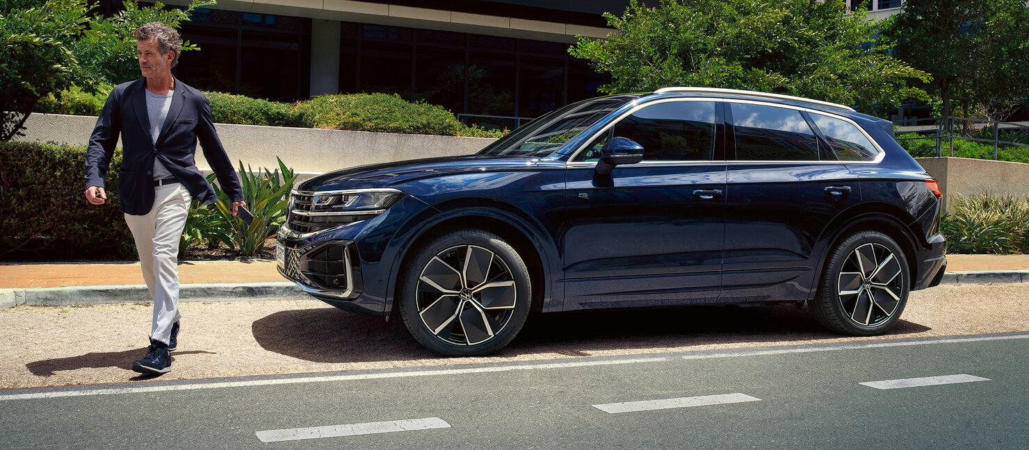 Dunkelblauer Volkswagen Touareg am Straßenrand, daneben ein Mann in Jacke und heller Hose vor Bürogebäude