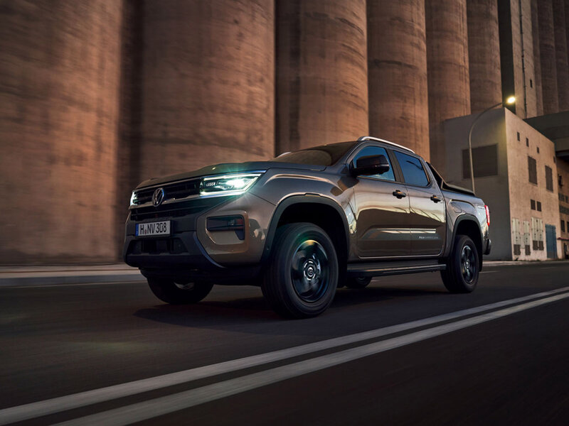 Ein Volkswagen Amarok fährt bei Dämmerung durch eine urbane Industrieumgebung. Das Fahrzeug ist frontal seitlich zu sehen, mit eingeschalteten LED-Scheinwerfern und erhöhter Bodenfreiheit.