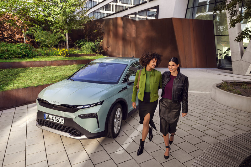Zwei Personen gehen an einem geparkten Škoda Elroq vor moderner Architektur vorbei.