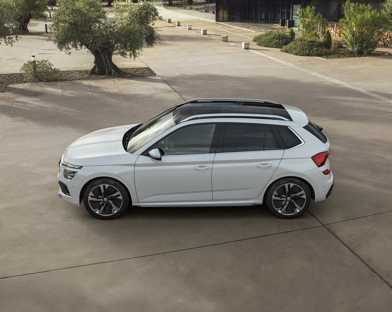 Škoda Kamiq in Weiß mit schwarzem Dach steht seitlich auf einem Platz vor moderner Architektur und Bäumen.