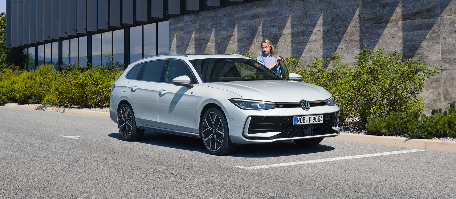 Weißer Volkswagen Passat Variant parkt vor modernem Gebäude, Person steht an offener Fahrertür