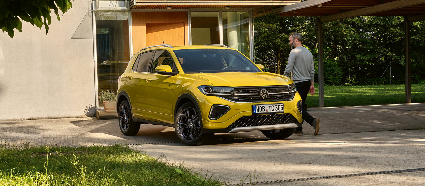 Gelber Volkswagen T-Cross steht vor modernem Haus, ein Mann geht daran vorbei – urbanes SUV im Alltagseinsatz