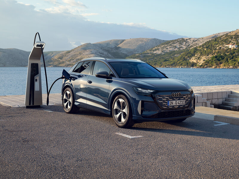 Audi Q4 e-tron steht seitlich an Ladesäule auf Parkplatz an einer Küstenstraße mit Bergen und Wasser im Hintergrund