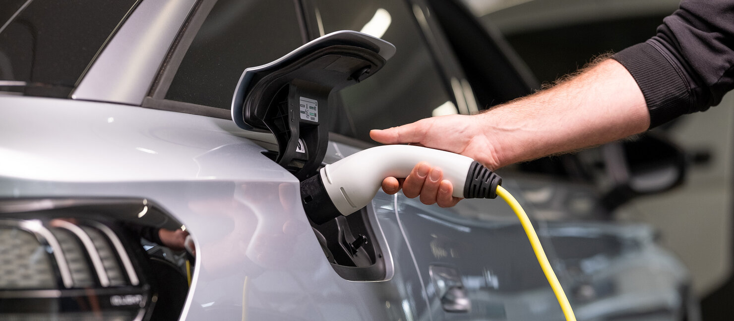 Eine Hand schließt ein gelbes Ladekabel an den Ladeanschluss eines silbernen Autos im Auto Bach Showroom an