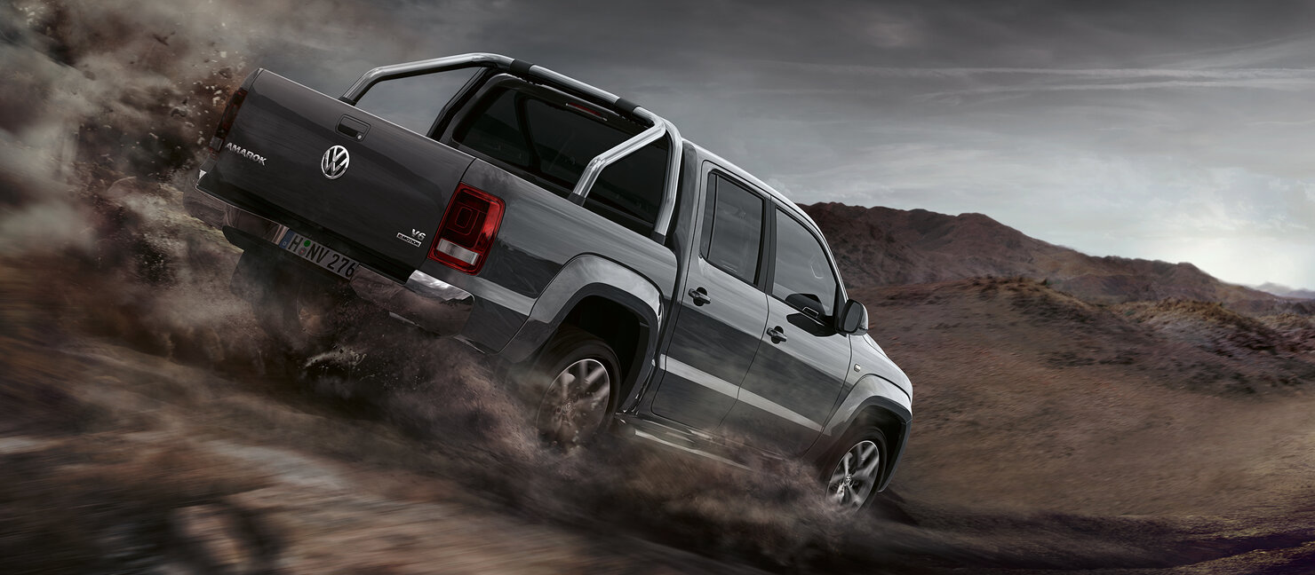 Grauer Volkswagen Amarok fährt durch felsiges Gelände, Staub wirbelt auf, raue Landschaft im Hintergrund.