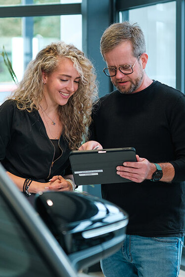 Zwei Mitarbeitende stehen im Autohaus am Fahrzeug und besprechen gemeinsam Informationen auf einem Tablet.