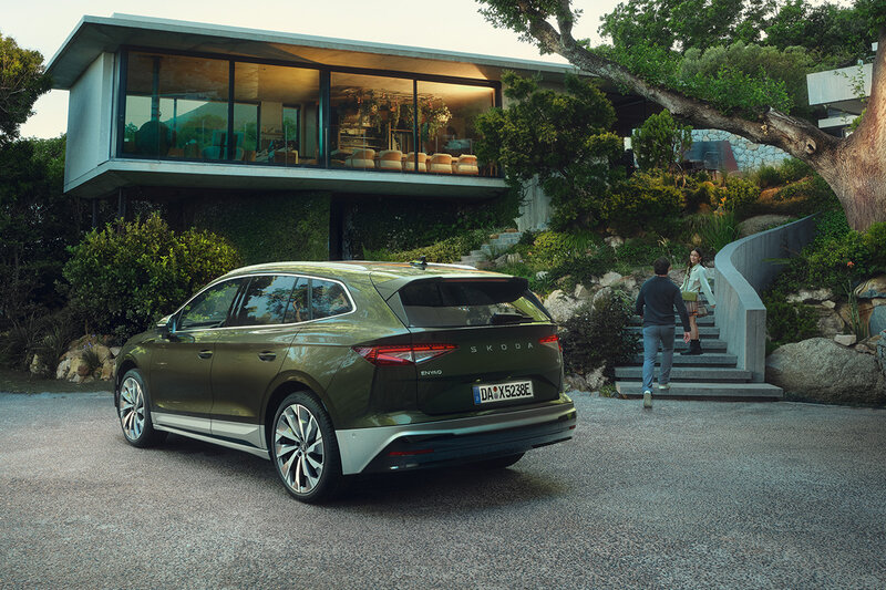 Škoda Enyaq in Olivgrün steht vor einem modernen Wohnhaus in grüner Umgebung bei Abendlicht.