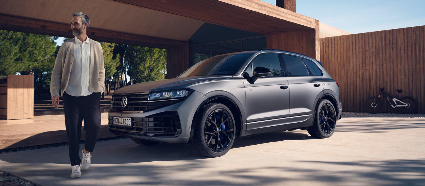 Grauer Volkswagen Touareg R eHybrid vor modernem Holzhaus, ein Mann geht daran vorbei, elegantes SUV-Design im Wohnumfeld