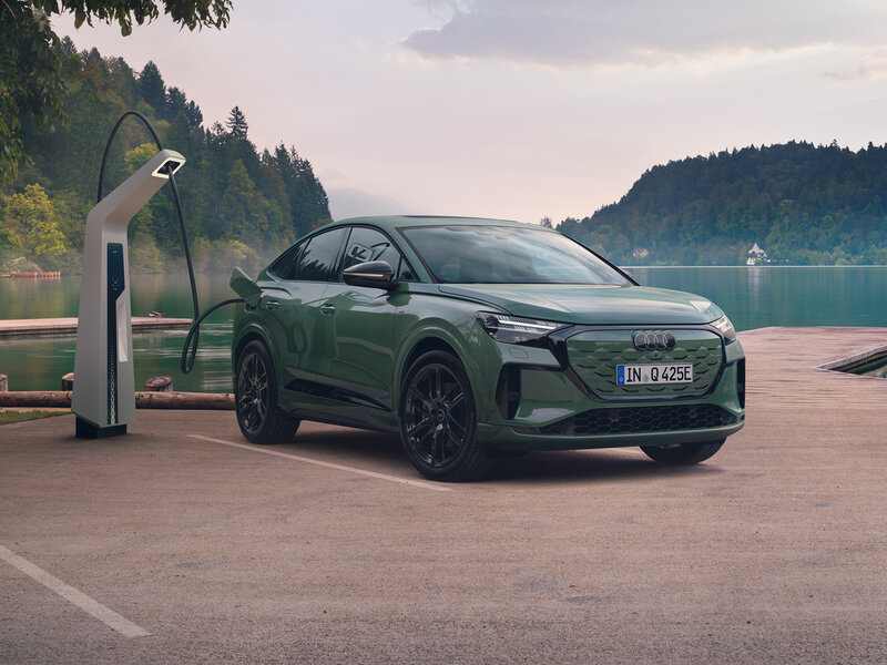Audi Q4 Sportback e-tron in Grün steht schräg vor Ladesäule auf Parkplatz an Seeufer mit bewaldeten Hügeln im Hintergrund