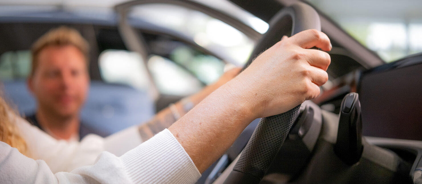Person hält das Lenkrad eines Fahrzeugs im Auto Bach Showroom, während ein Mitarbeiter neben dem Auto hockt