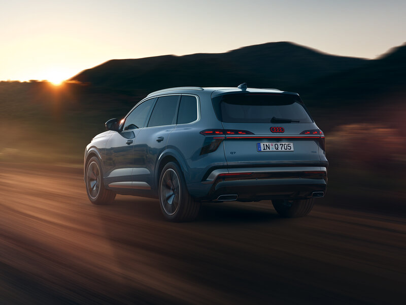 Audi Q7 in Heckansicht fährt auf einer Landstraße bei Sonnenuntergang vor bergiger Landschaft mit Bewegungsunschärfe im Hintergrund
