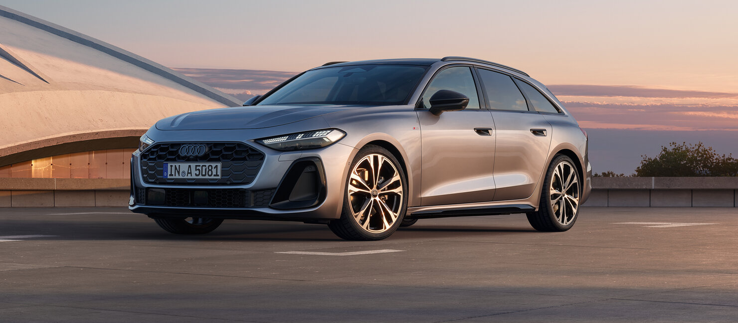 Silberner Audi A5 Avant in frontaler Schrägansicht auf weitläufiger Fläche vor moderner Architektur bei Abendlicht