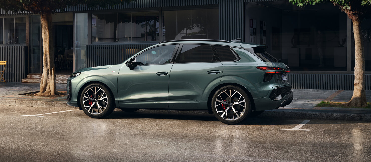 Audi Q3 in Seitenansicht vor urbaner Kulisse mit Bäumen, dynamische Linienführung und erhöhte Bodenfreiheit sichtbar