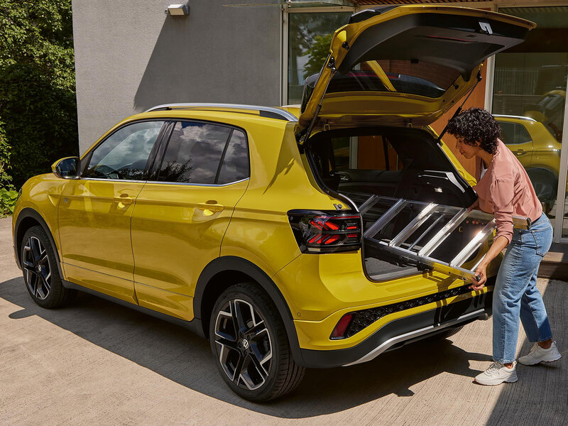 Offener Kofferraum eines gelben Volkswagen T-Cross, während eine Person eine zusammengeklappte Leiter einlädt.