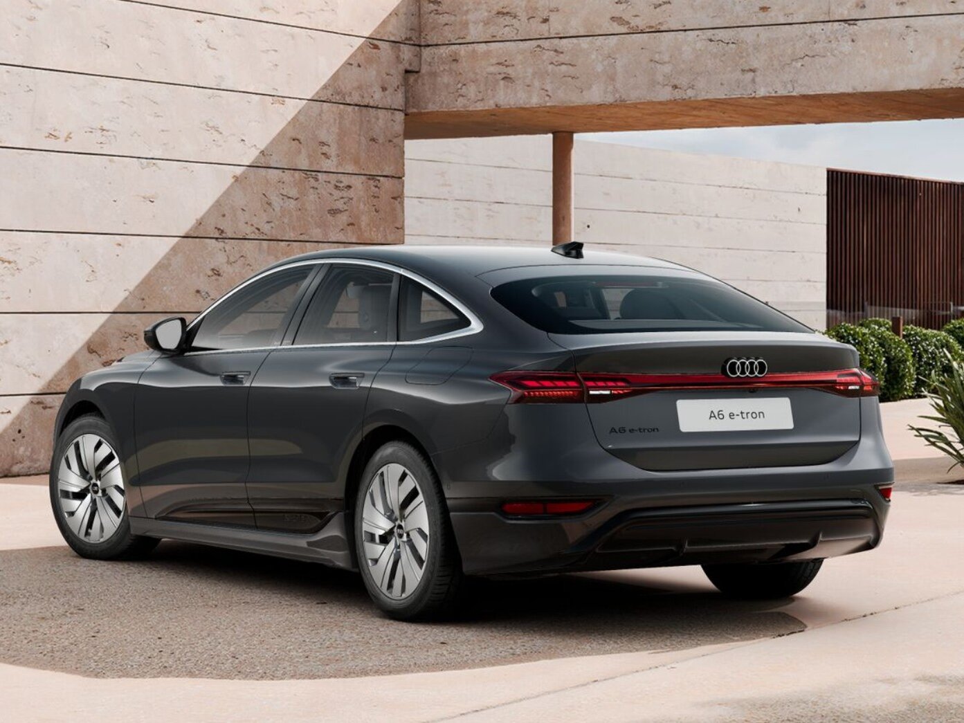 Dunkelgrauer Audi A6 e-tron in Heckansicht steht vor moderner Architektur mit Steinfassade und begrüntem Außenbereich