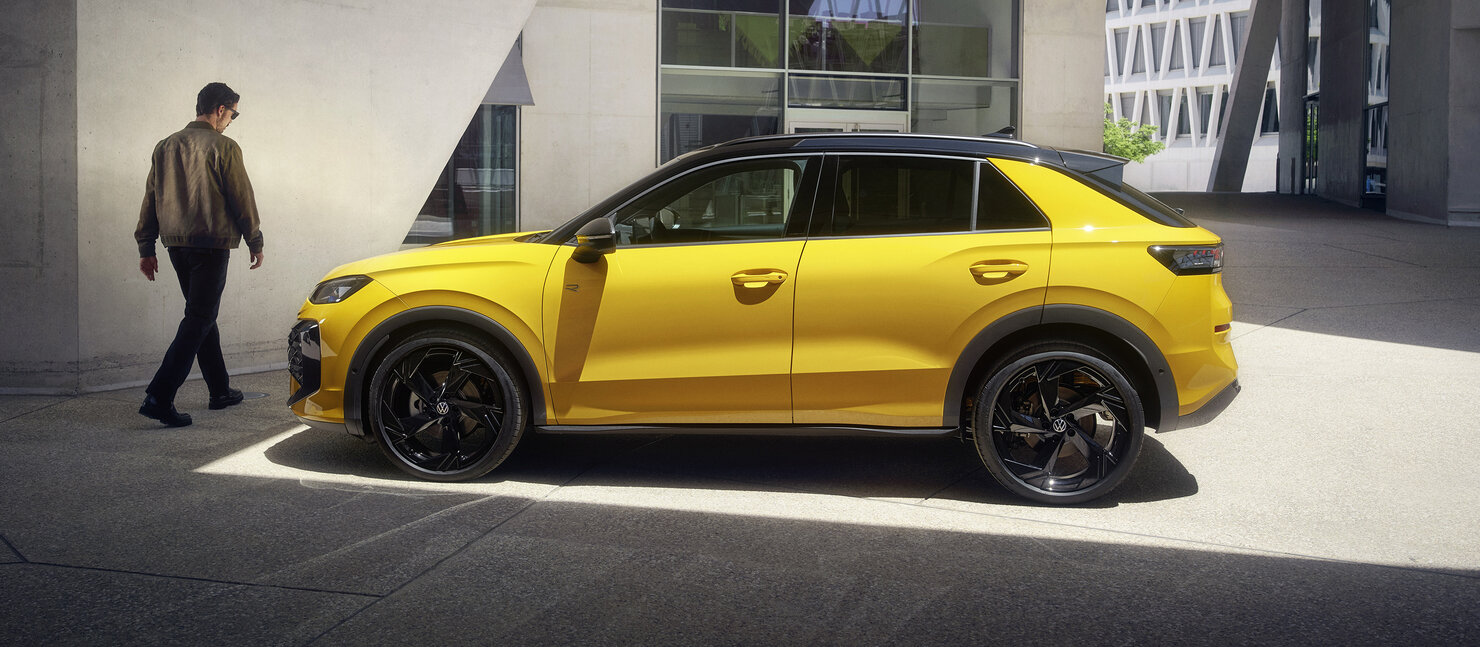 Gelber Volkswagen T-Roc in Seitenansicht, geparkt vor modernem Gebäude, eine Person geht am Fahrzeug vorbei