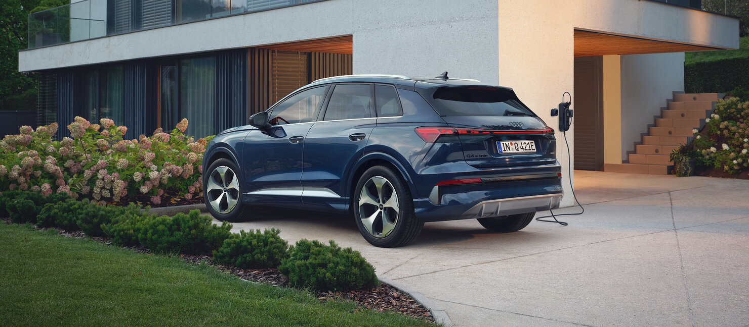 Audi Q4 e-tron in Blau lädt an Wallbox vor modernem Einfamilienhaus aus Heckperspektive mit Garten und Einfahrt