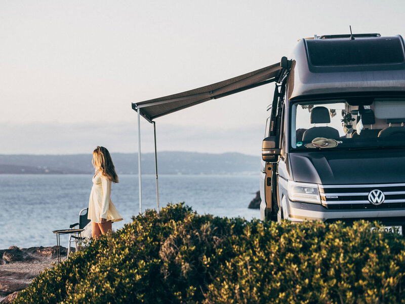 Ein Volkswagen Grand California steht an einer Küste mit ausgefahrener Markise. Eine Person steht vor dem Fahrzeug und blickt auf das Wasser. Der Fokus liegt auf Reisemobil-Design und Outdoor-Nutzung.
