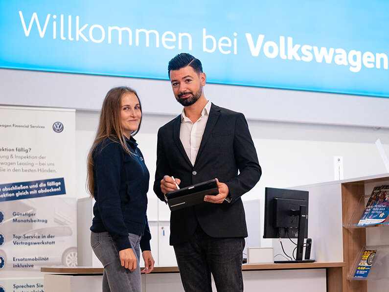 Zwei Personen stehen im Volkswagen Autohaus Auto Bach vor einem Empfangsbereich mit Tablet und Willkommensschild im Hintergrund