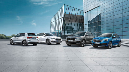 Škoda Modelle stehen auf einem Platz vor moderner Glasarchitektur, darunter Kompaktwagen, Limousine, SUV und Kombi aus verschiedenen Fahrzeugklassen.