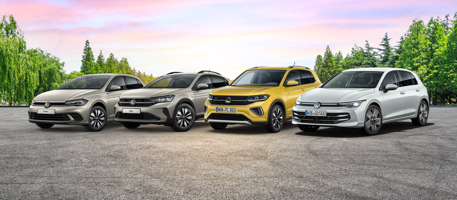 Vier Volkswagen Modelle stehen nebeneinander auf einer asphaltierten Fläche vor einer Baumreihe bei Sonnenuntergang