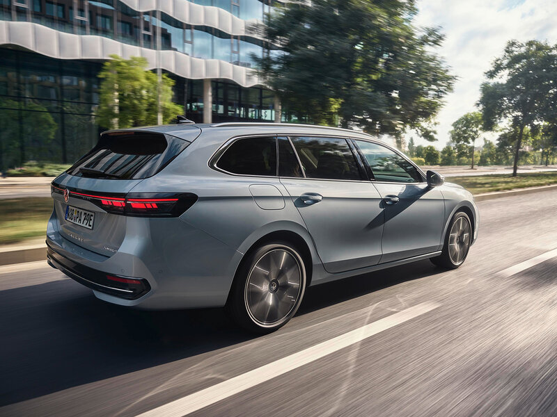 Volkswagen Passat eHybrid in Silber fährt auf einer städtischen Straße bei Tageslicht vorbei an moderner Glasarchitektur und Grünflächen