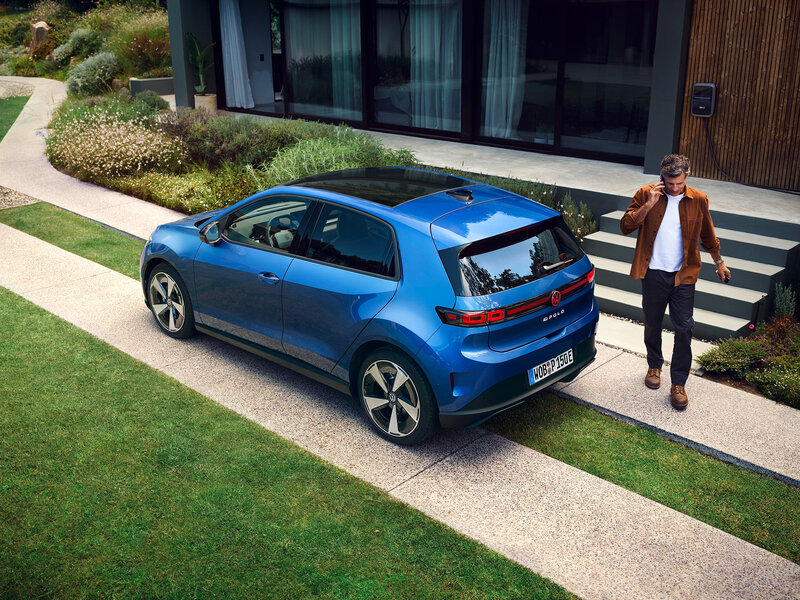 Volkswagen ID. Polo in Blau in Heck-Dreiviertelansicht steht auf einer Einfahrt vor modernem Wohnhaus mit Person neben dem Fahrzeug
