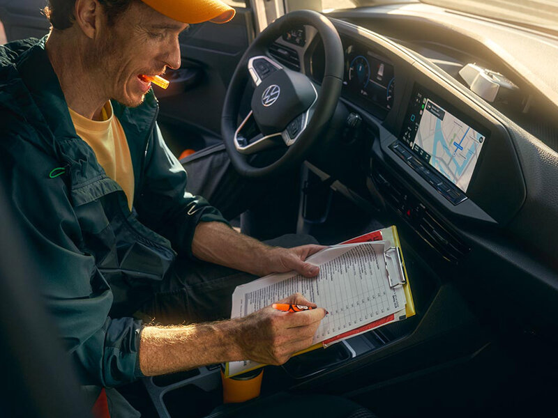 Blick in den Fahrerraum des Volkswagen Caddy Cargo. Eine Person sitzt auf dem Fahrersitz und nutzt Unterlagen, das Cockpit mit Lenkrad und Navigationsdisplay ist sichtbar.