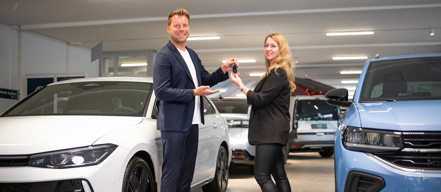 Zwei Personen stehen im Volkswagen Autohaus Auto Bach zwischen Fahrzeugen, während eine Autoschlüsselübergabe zwischen einem weißen und hellblauen Volkswagen stattfindet
