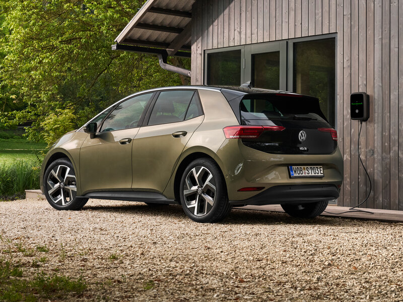 Ein kompakter Volkswagen ID.3 steht vor einem Holzhaus, seitlich von hinten gezeigt, mit Ladeanschluss an einer Wallbox.