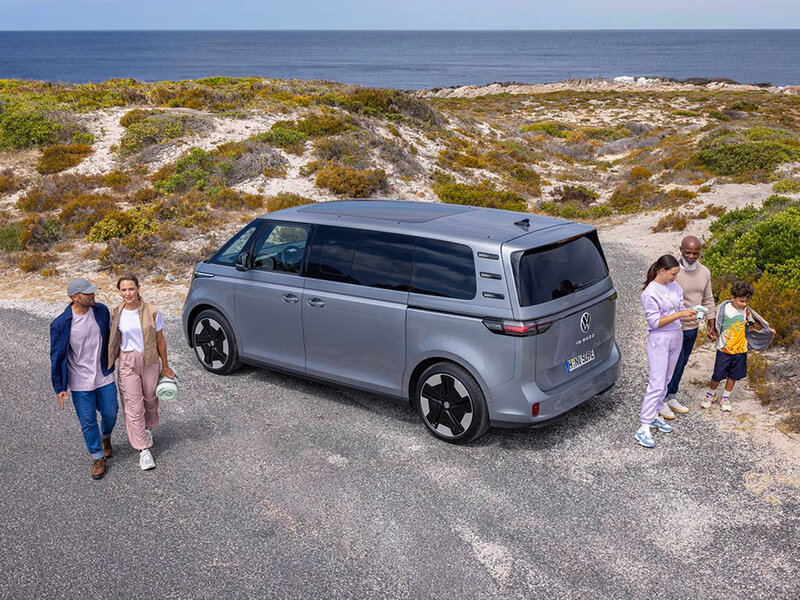 Ein grauer Volkswagen ID. Buzz steht auf einem Parkplatz in Küstennähe. Mehrere Personen gehen neben dem Fahrzeug, die Umgebung ist offen und naturnah.
