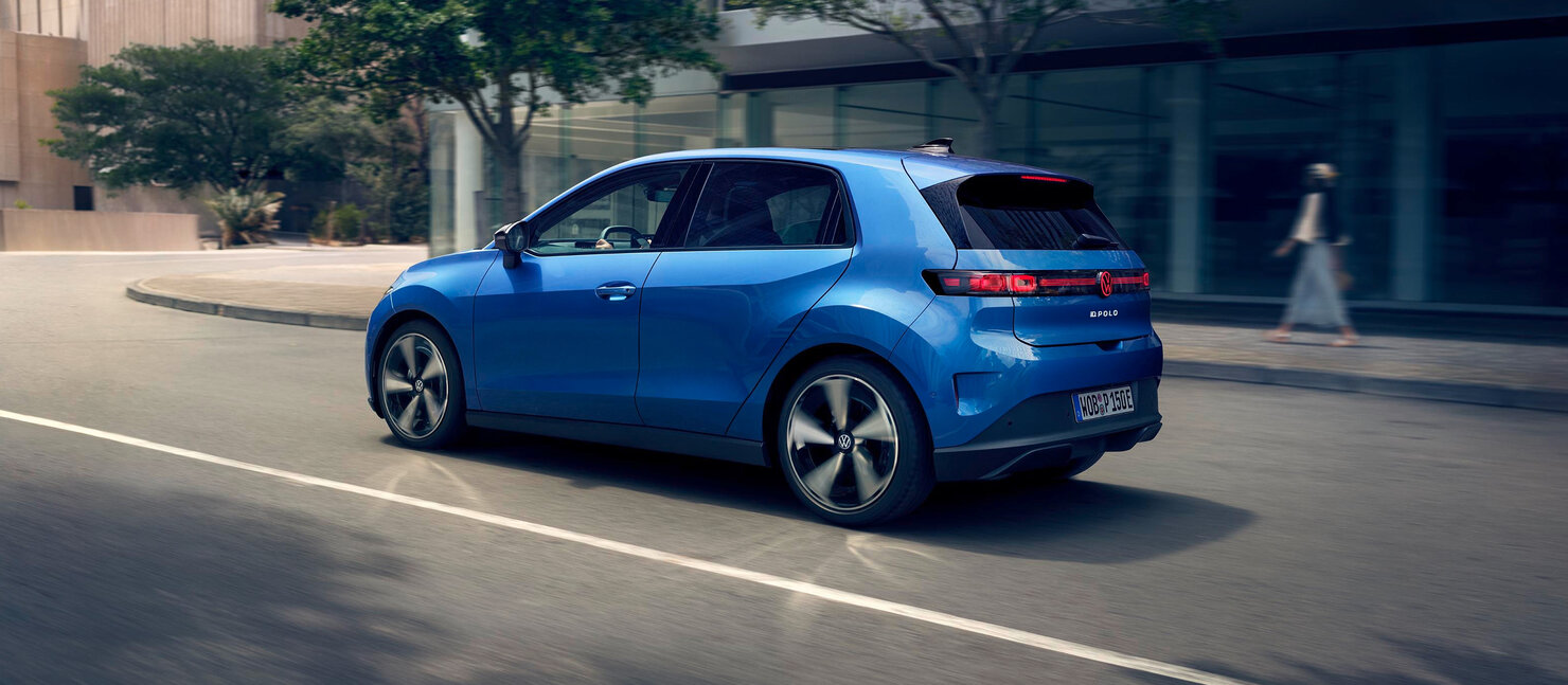 Volkswagen ID. Polo in Blau in Heck-Dreiviertelansicht fährt auf einer Stadtstraße vor moderner Glasfassade