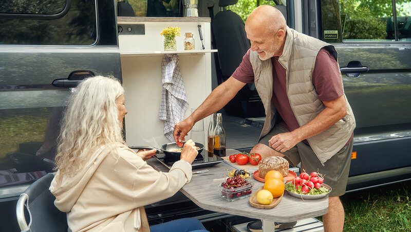 Zwei Personen bereiten eine Mahlzeit an der Außenküche eines Volkswagen Grand California vor