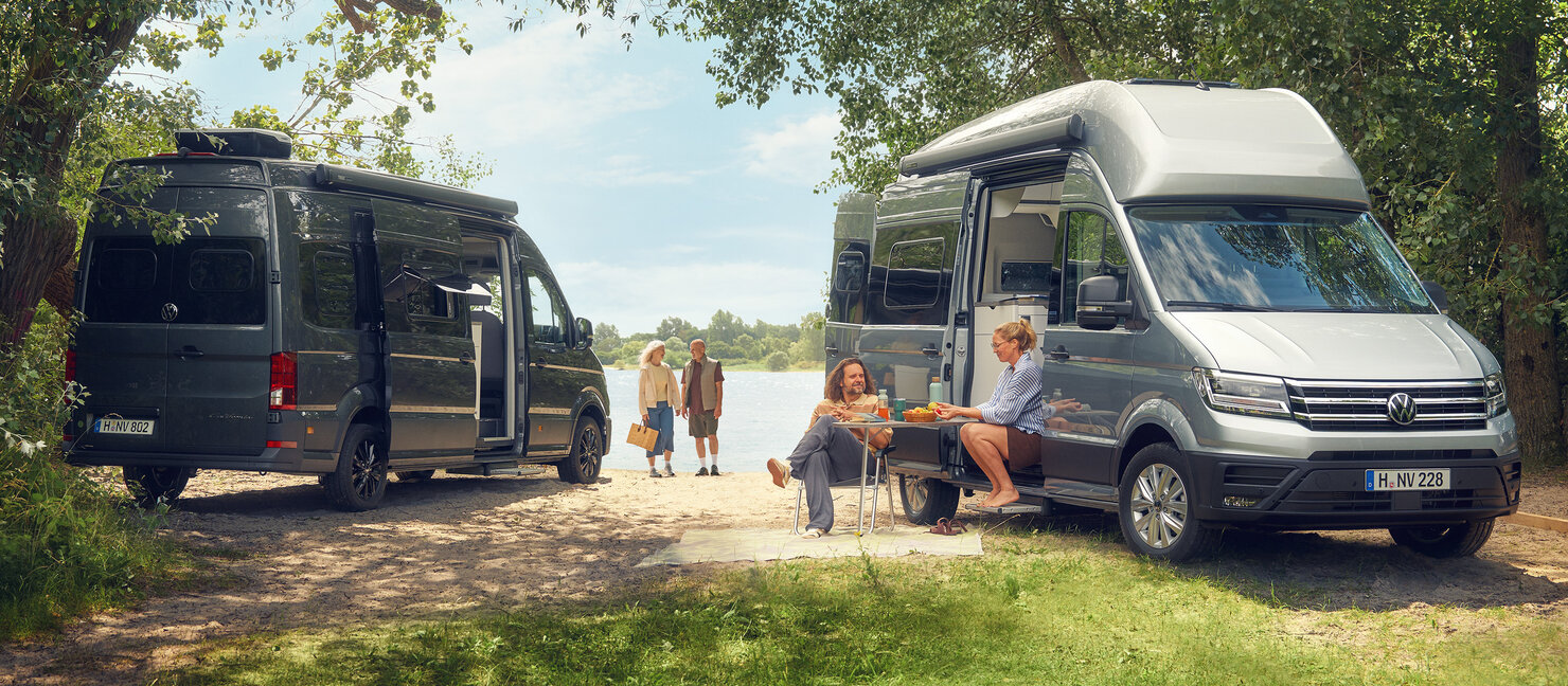 Zwei Volkswagen Grand California stehen am Seeufer unter Bäumen, während Personen am Wasser entspannen