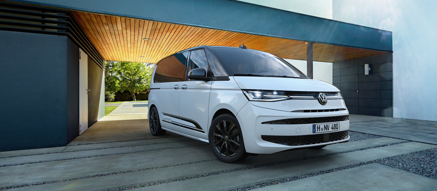 Weißer Volkswagen Multivan steht vor modernem Holzgebäude, klare Architektur und Tageslicht.