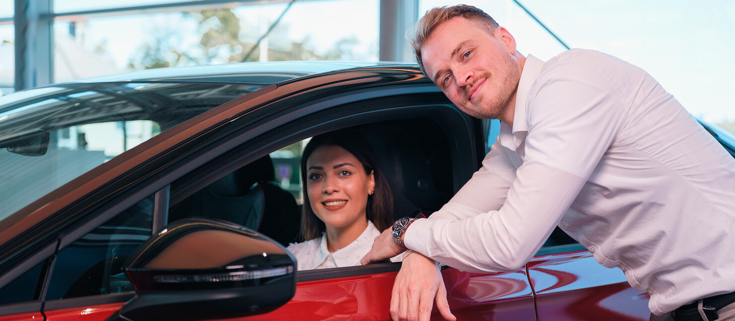 Eine Frau sitzt in einem roten Auto, während ein Auto Bach Mitarbeiter ihr im Showroom eine Fahrzeugfunktion erklärt