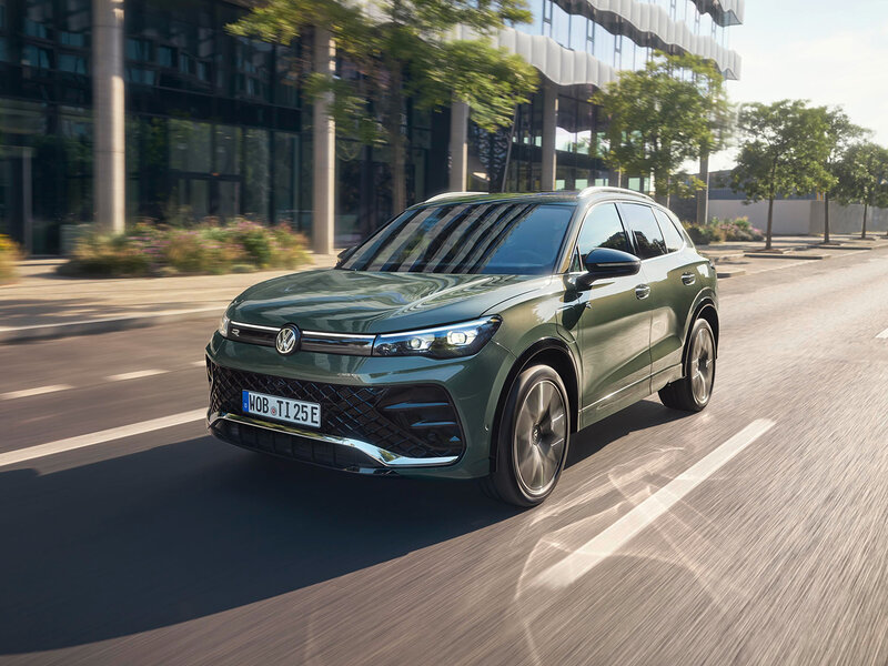 Volkswagen Tiguan eHybrid in Dunkelgrün fährt auf einer städtischen Straße vor moderner Glasfassade bei Tageslicht
