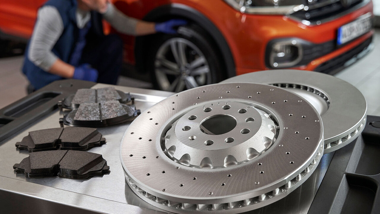 Bremsscheiben und Bremsbeläge liegen auf einem Werkstatttisch, im Hintergrund ein Mechaniker bei der Wartung eines orangefarbenen Volkswagen SUV