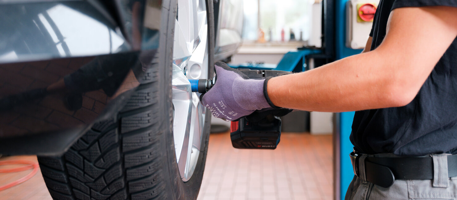 Person montiert Rad an Fahrzeug mit Akkuschrauber in Werkstattumgebung, Nahaufnahme auf Reifen und Radschrauben während Wartungsarbeit