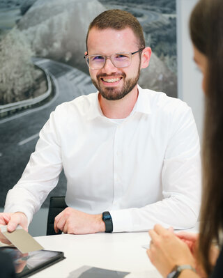 Lächelnder Autohaus-Mitarbeiter im Beratungsgespräch mit Kundinnen, am Tisch sitzend und Musterkarten erklärend.